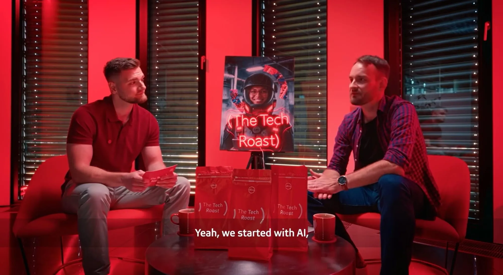 Two men sitting on red sofas on a red-lit studio set, talking across a low table holding branded coffee bags and mugs, with a framed show poster reading "The Tech Roast" between them and a burned-in subtitle reading "Yeah, we started with AI,"