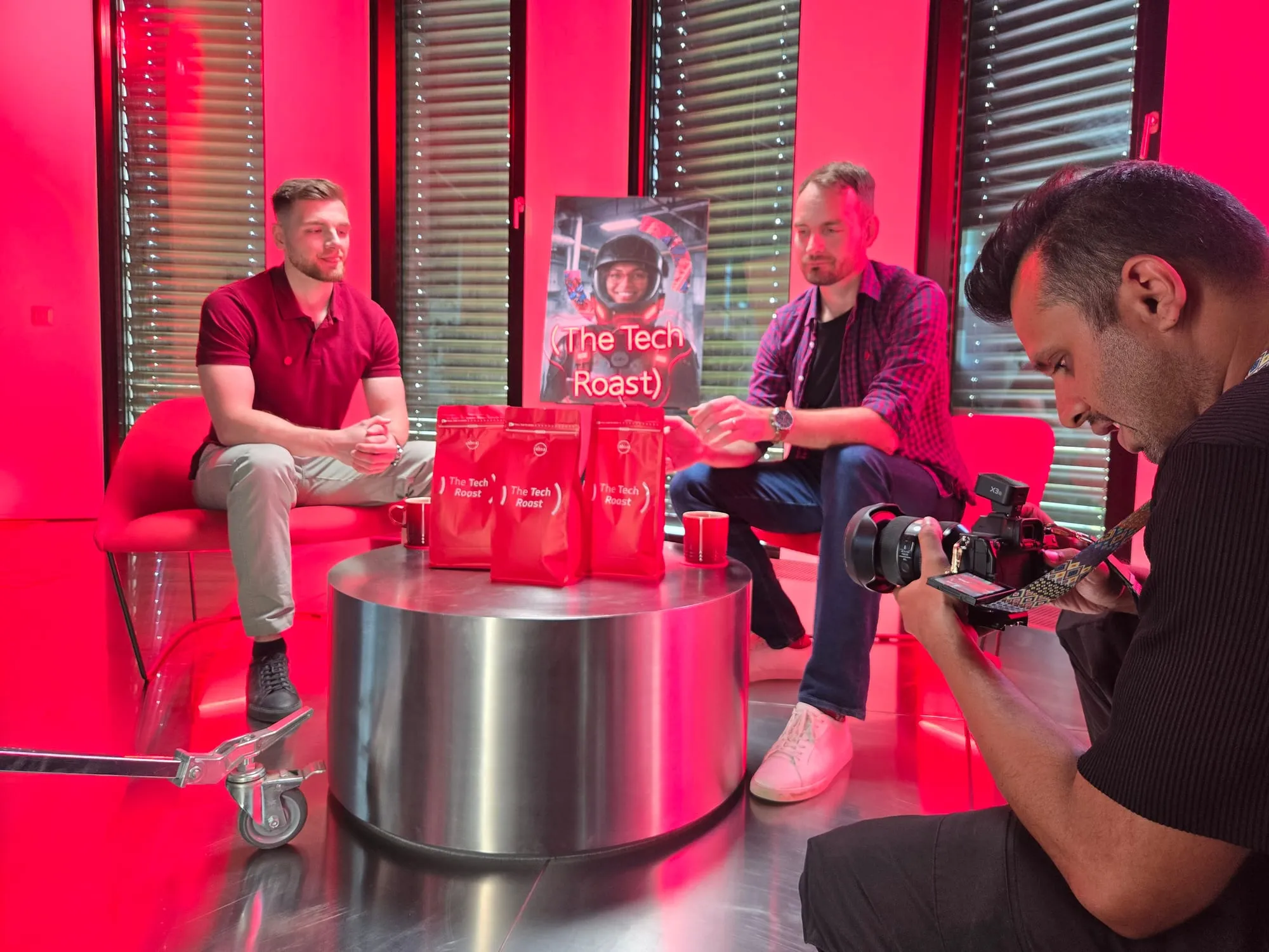Two people talking on the studio set, with branded coffee bags arranged on a low metal table between them and a photographer shooting in the foreground