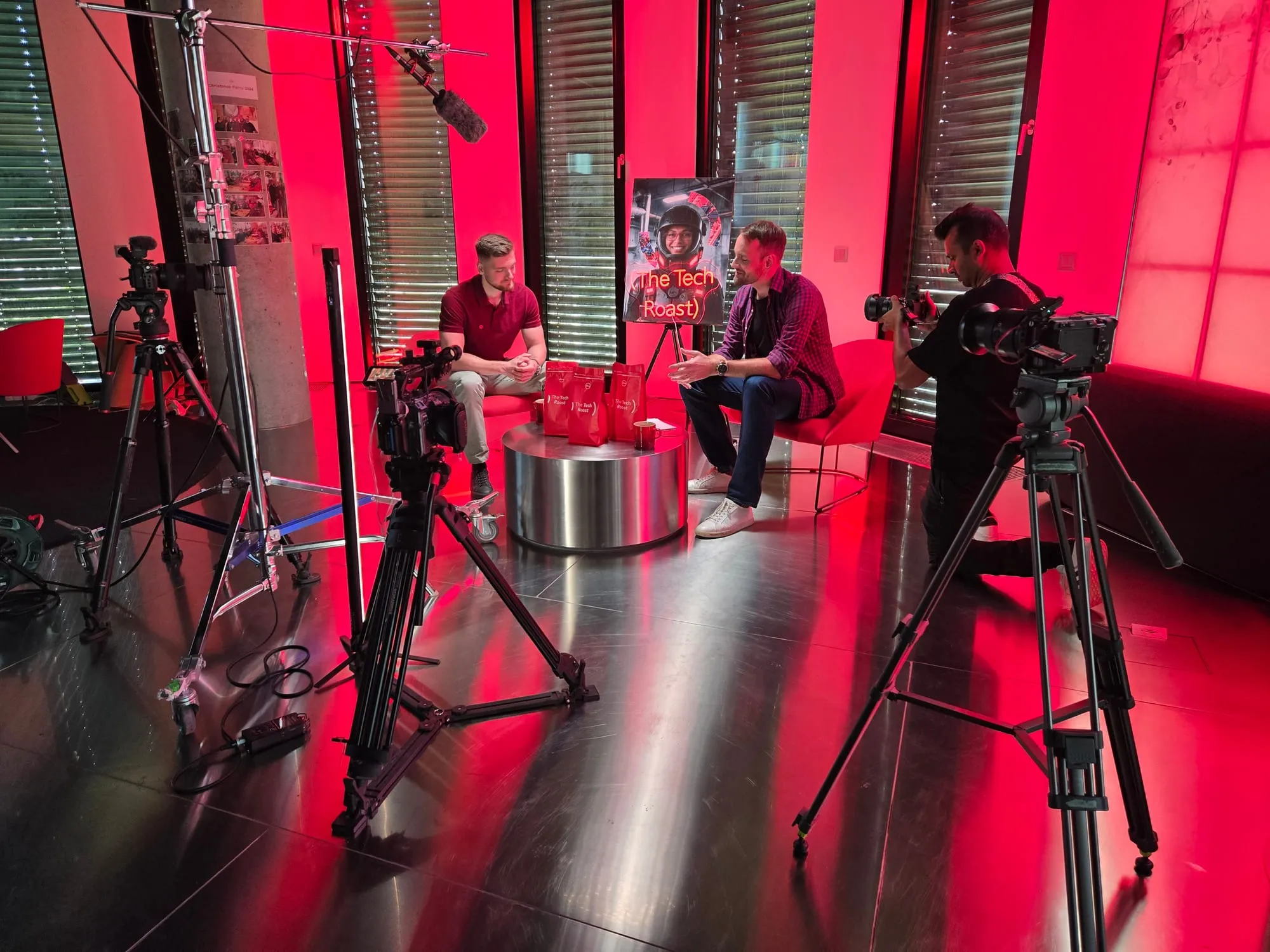 A red-lit studio set with two people seated in conversation, surrounded by cameras on tripods and a boom microphone